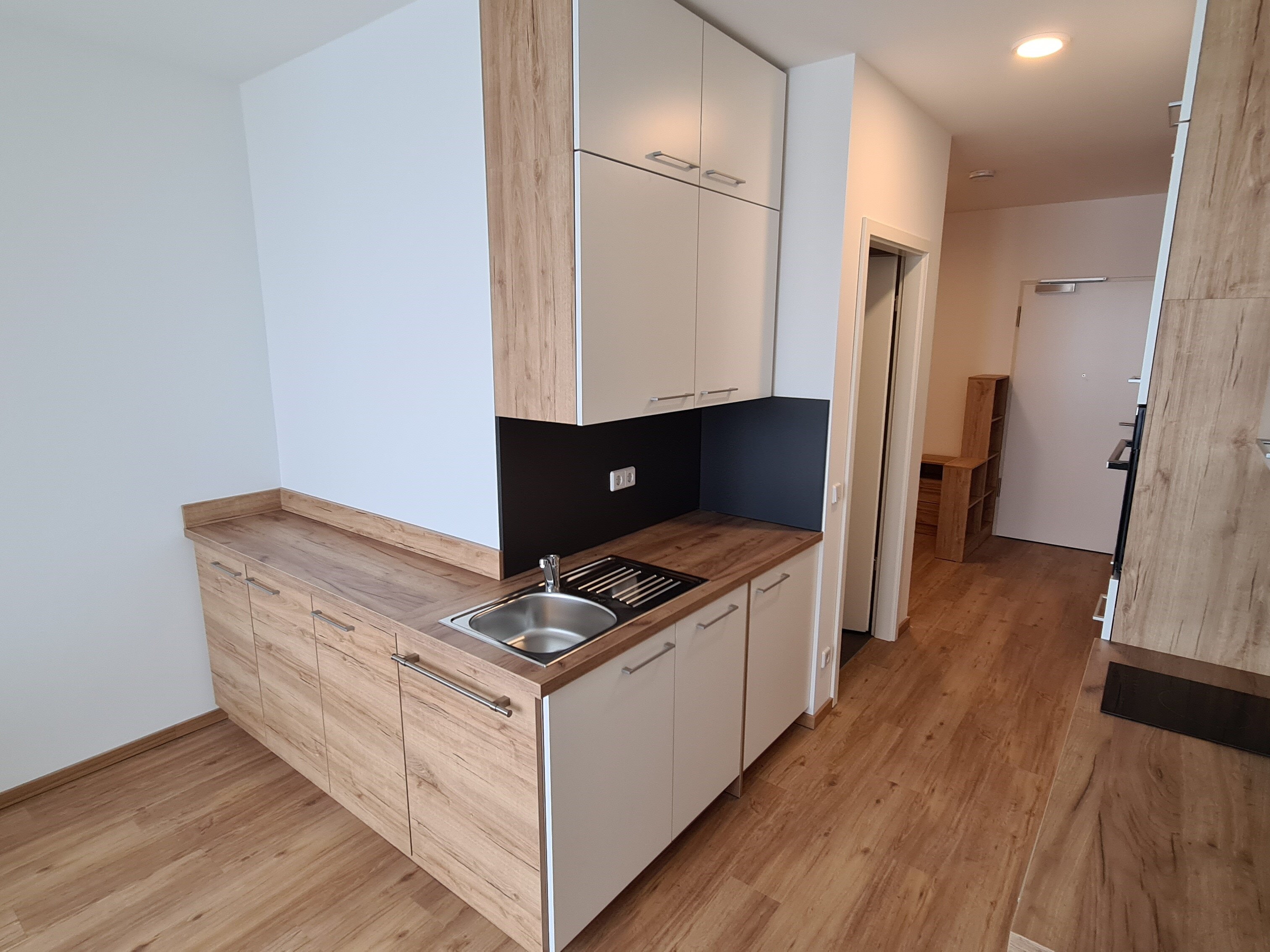 Schmale Einbauküche mit hellen Fronten und Holzoptik-Arbeitsplatte, ausgestattet mit Spüle, Kochfeld und mehreren Ober- und Unterschränken; im Hintergrund ist der Flur mit Wohnungseingangstür sichtbar.