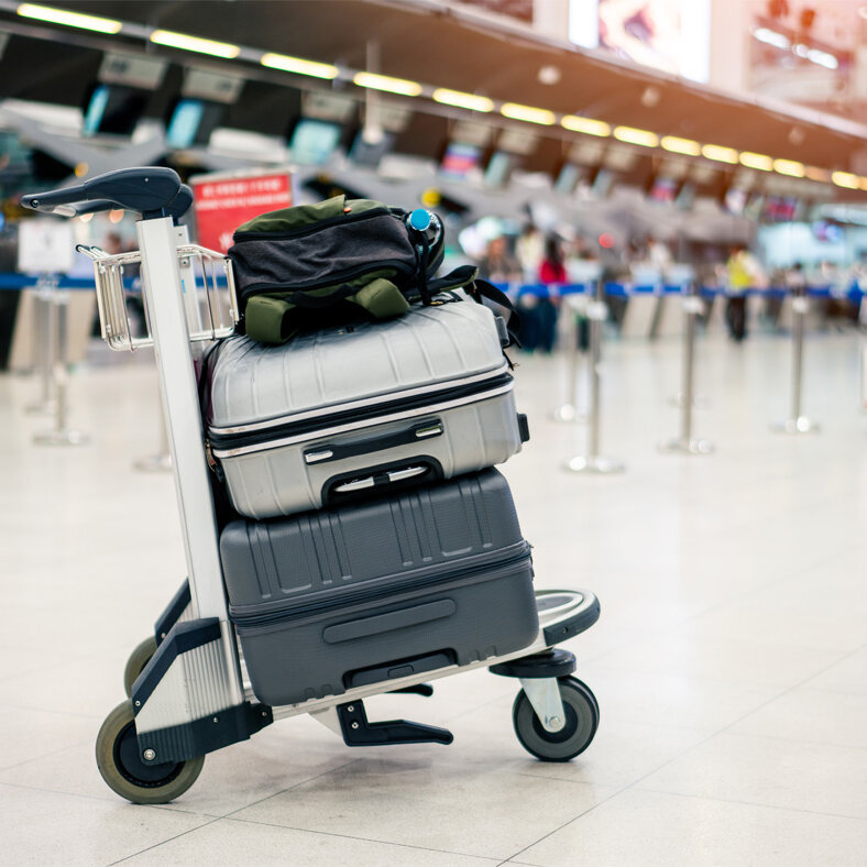 Gepäckwagen mit mehreren Koffern steht in einer Flughafenhalle vor Check-in-Schaltern; im Hintergrund befinden sich Absperrbänder und wartende Reisende.