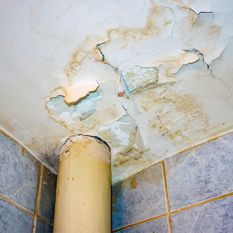 Decke eines Badezimmers mit sichtbaren Feuchtigkeitsflecken, abblätternder Farbe und Rissen rund um ein Rohr; darunter sind geflieste Wände zu sehen.