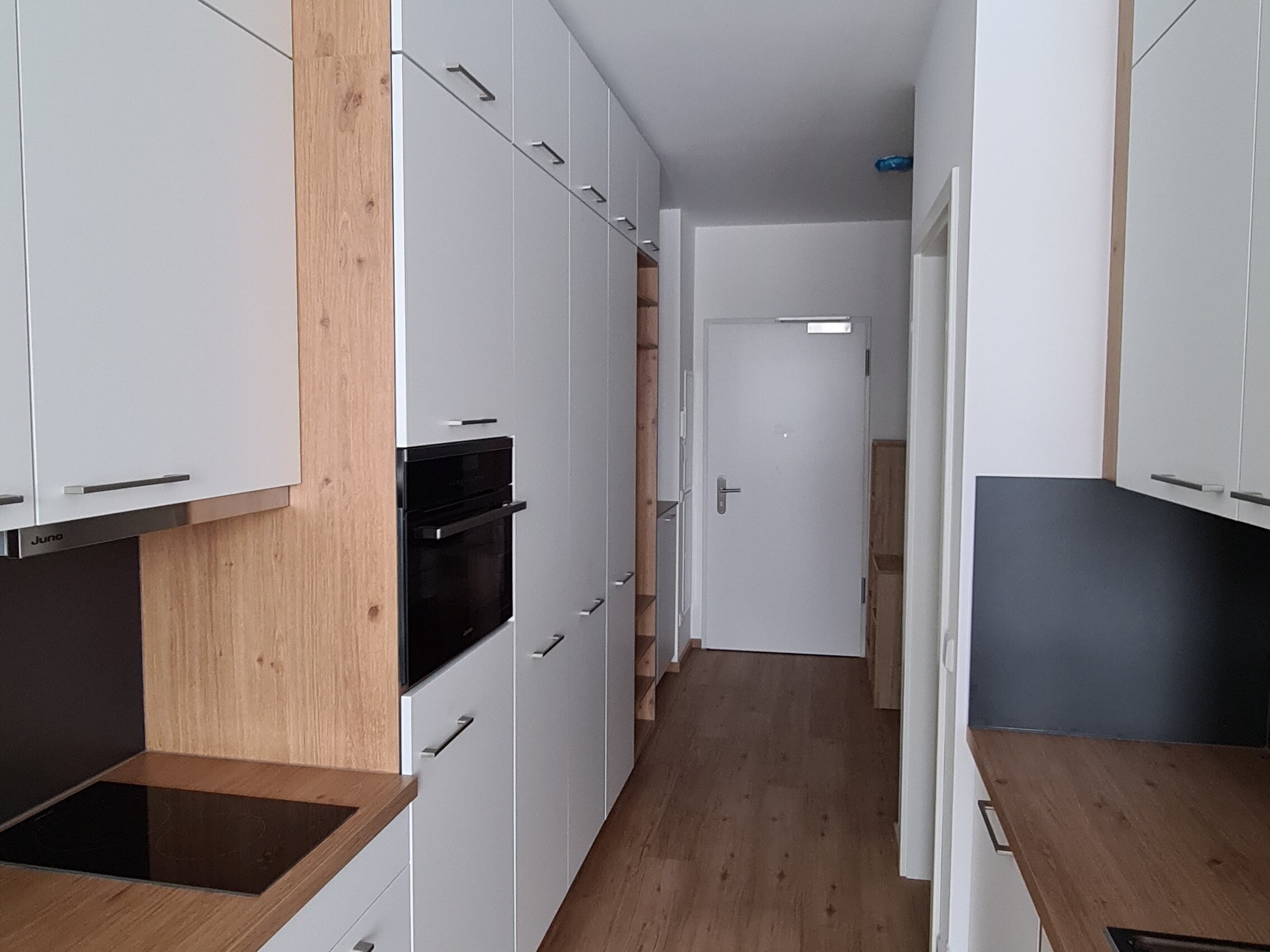 Lange, schmale Küchenzeile mit beidseitigen Einbauschränken in Weiß und Holzoptik, integriertem Backofen und Kochfeld; am Ende des Flurs befindet sich die Wohnungseingangstür.