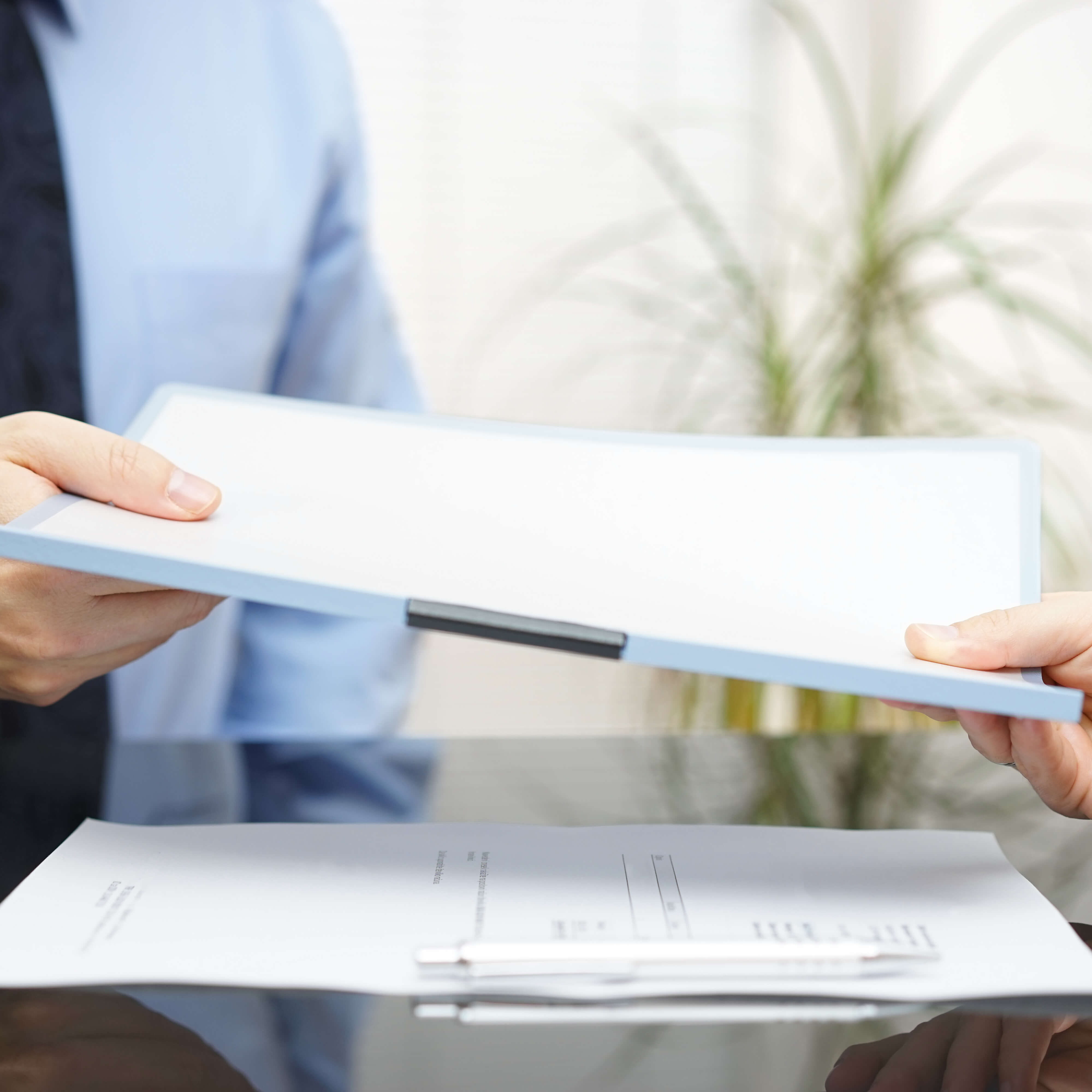 Zwei Personen übergeben sich an einem Tisch einen geschlossenen Aktenordner; im Vordergrund liegen Dokumente, im Hintergrund ist eine Pflanze in einem hellen Büro zu sehen.