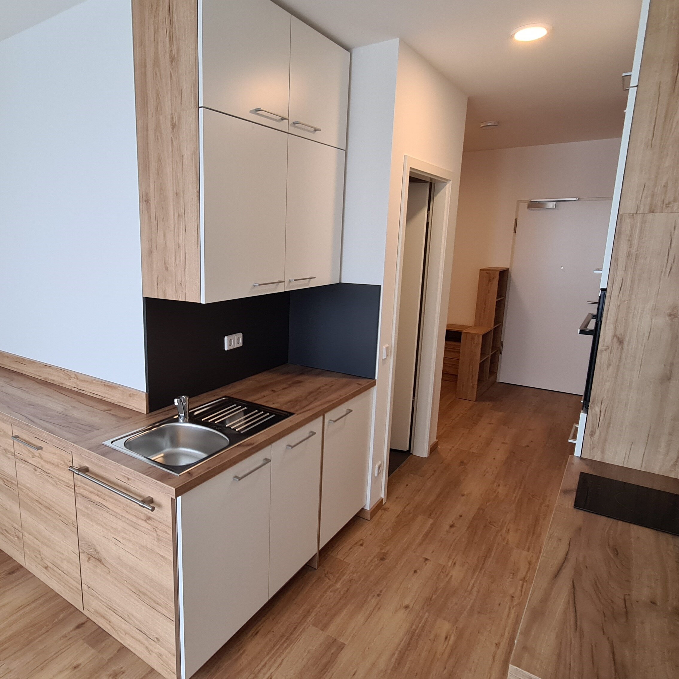 Schmale Einbauküche mit hellen Fronten und Holzoptik-Arbeitsplatte, ausgestattet mit Spüle, Kochfeld und mehreren Ober- und Unterschränken; im Hintergrund ist der Flur mit Wohnungseingangstür sichtbar.