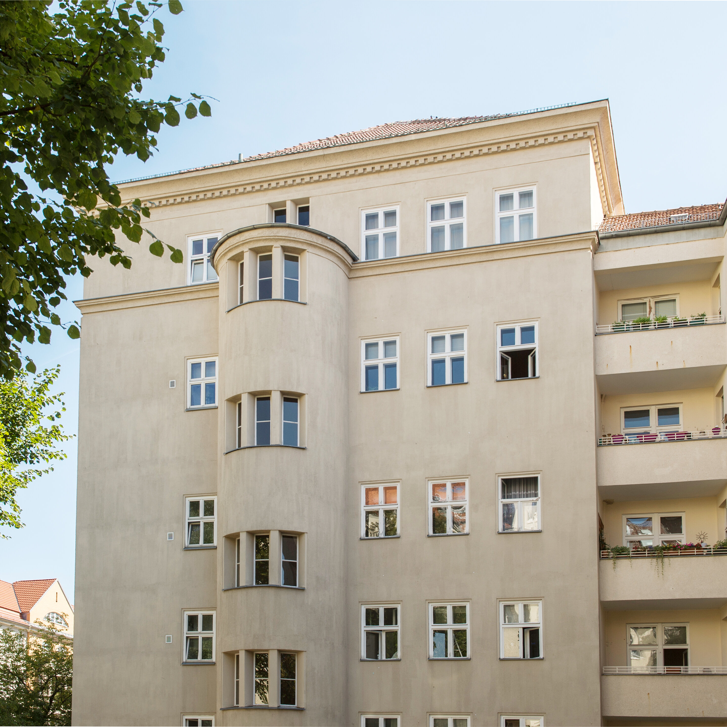 Mehrgeschossiges Wohngebäude mit heller Putzfassade, symmetrisch angeordneten Fenstern und einem halbrunden Erker in der Mitte; rechts befinden sich Balkone mit Geländern, links rahmen Bäume das Gebäude ein.