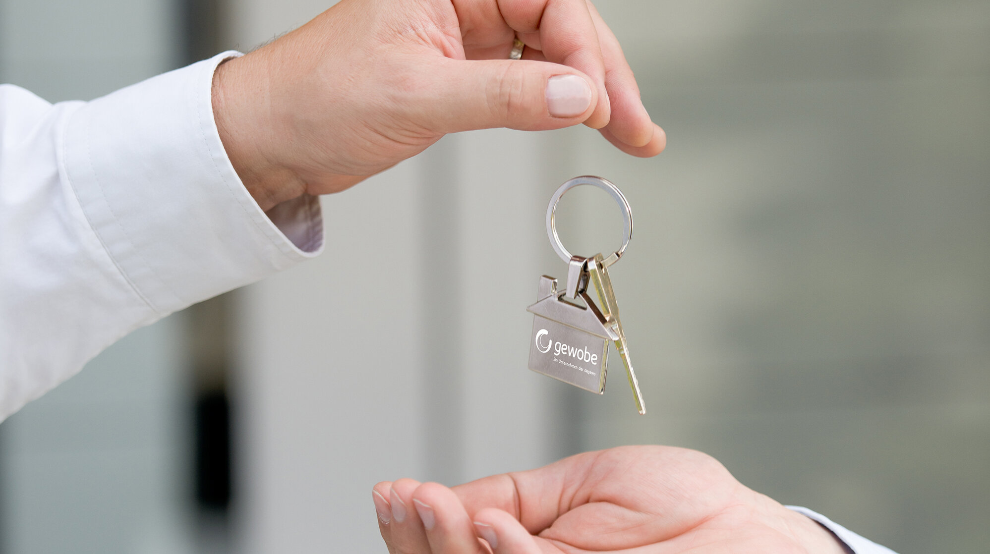 Eine Person übergibt einen Schlüsselbund mit zwei Schlüsseln und einem hausförmigen Anhänger mit „gewobe“-Logo an eine andere Person; die Hände sind im Vordergrund zu sehen, der Hintergrund ist unscharf und hell.