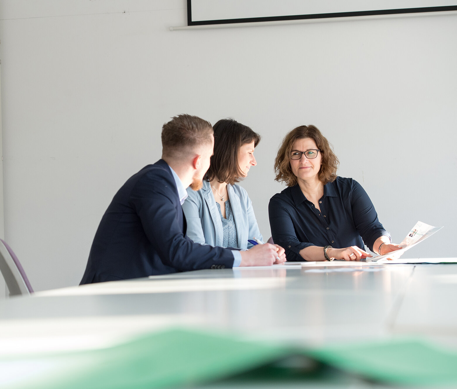 Drei Personen sitzen an einem Konferenztisch in einem hellen Raum und besprechen gemeinsam ein Dokument; eine Frau hält Unterlagen in der Hand, während die beiden anderen aufmerksam zuhören.