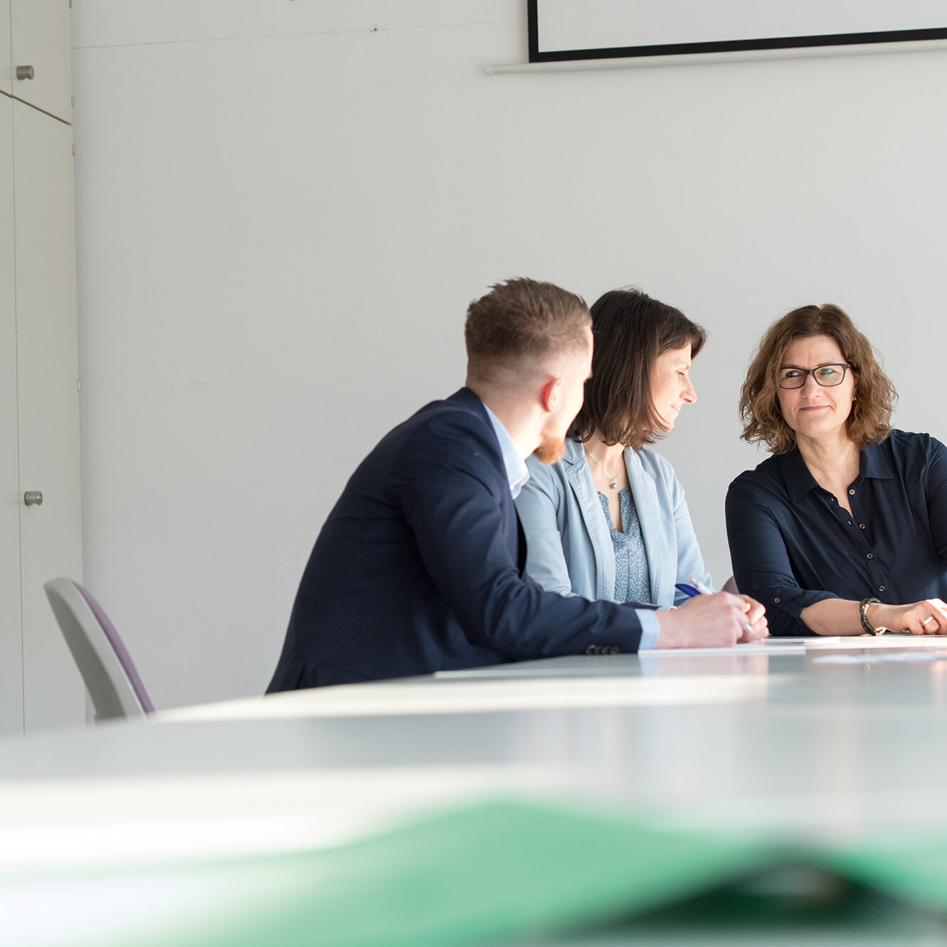 Drei Personen sitzen an einem Konferenztisch in einem hellen Raum und besprechen gemeinsam ein Dokument; eine Frau hält Unterlagen in der Hand, während die beiden anderen aufmerksam zuhören.
