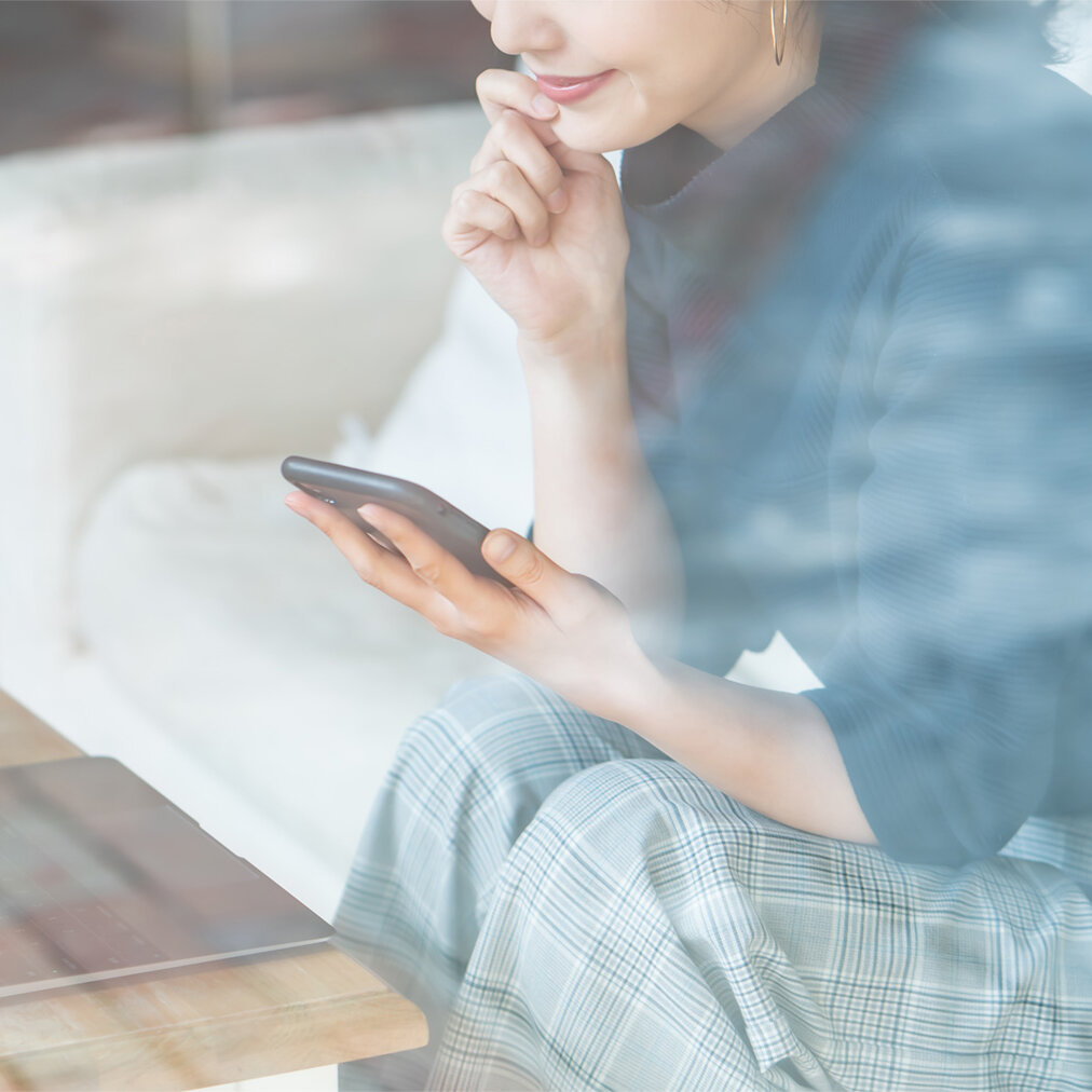 Eine Person sitzt auf einem Sofa und hält ein Smartphone in der Hand; auf einem Tisch davor stehen ein geöffnetes Laptop und ein Glas Getränk, im Hintergrund ist ein Fahrrad zu sehen.