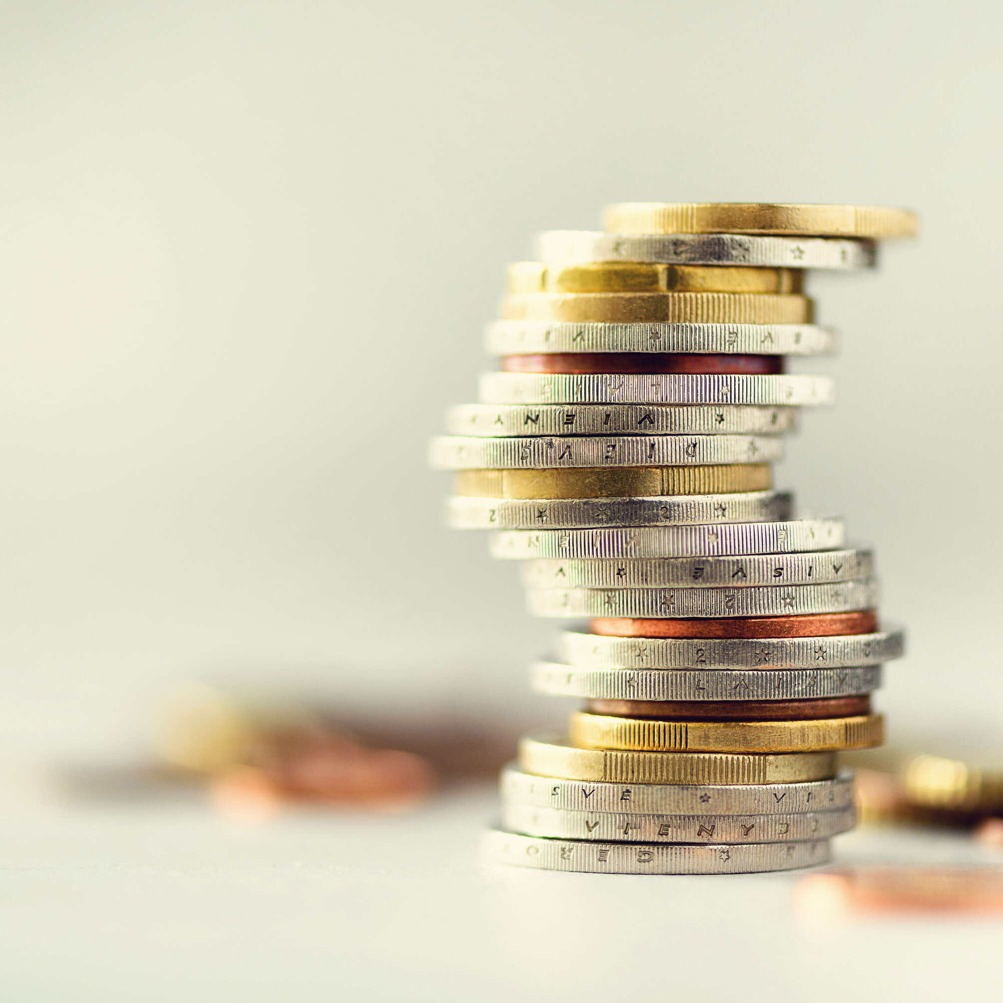 Mehrere übereinandergestapelte Euro-Münzen stehen auf einer hellen Oberfläche; im Hintergrund liegen weitere Münzen unscharf im Bild.