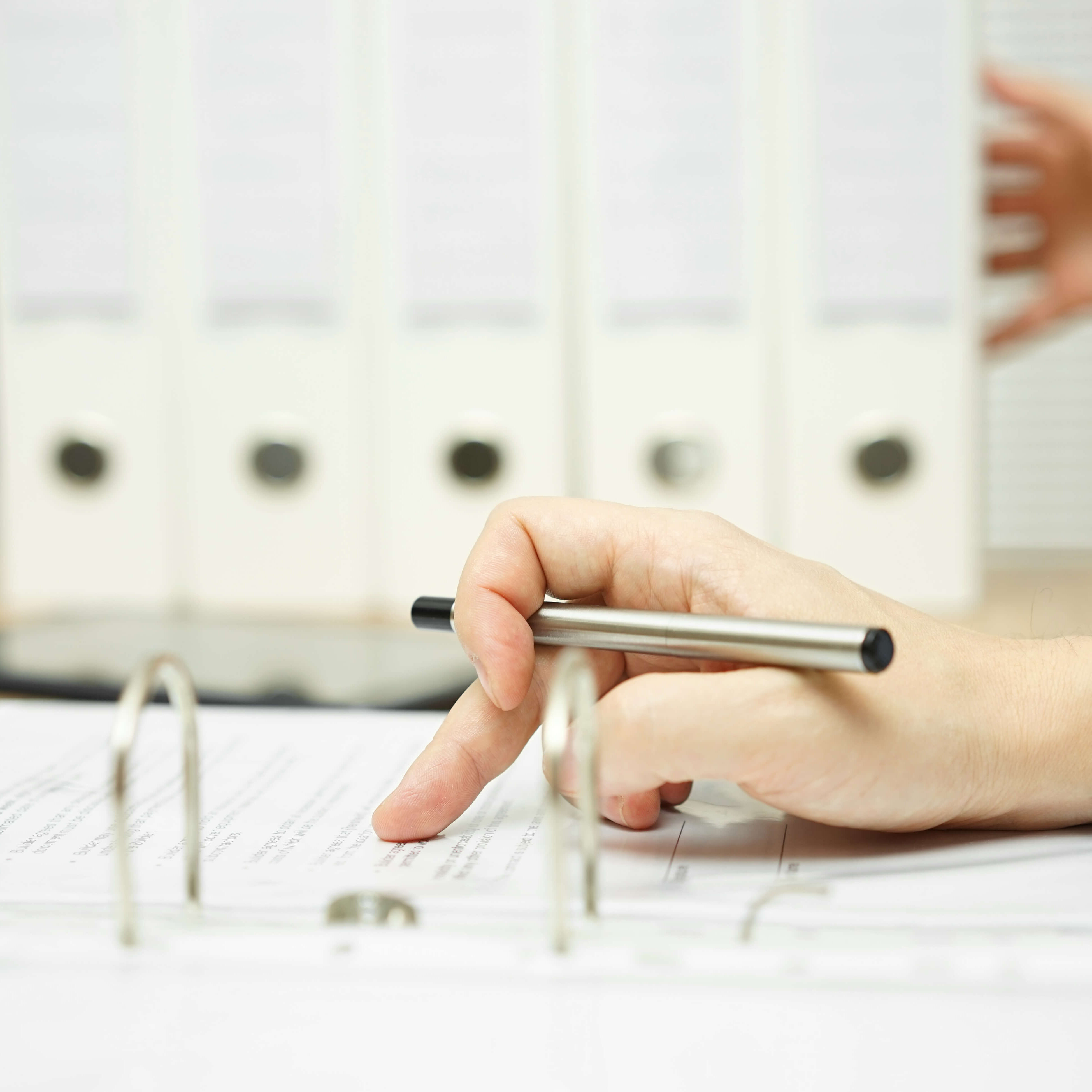 Eine Person blättert in einem geöffneten Ringordner und zeigt mit einem Stift auf ein Dokument; im Hintergrund stehen mehrere weiße Aktenordner in einem Regal.