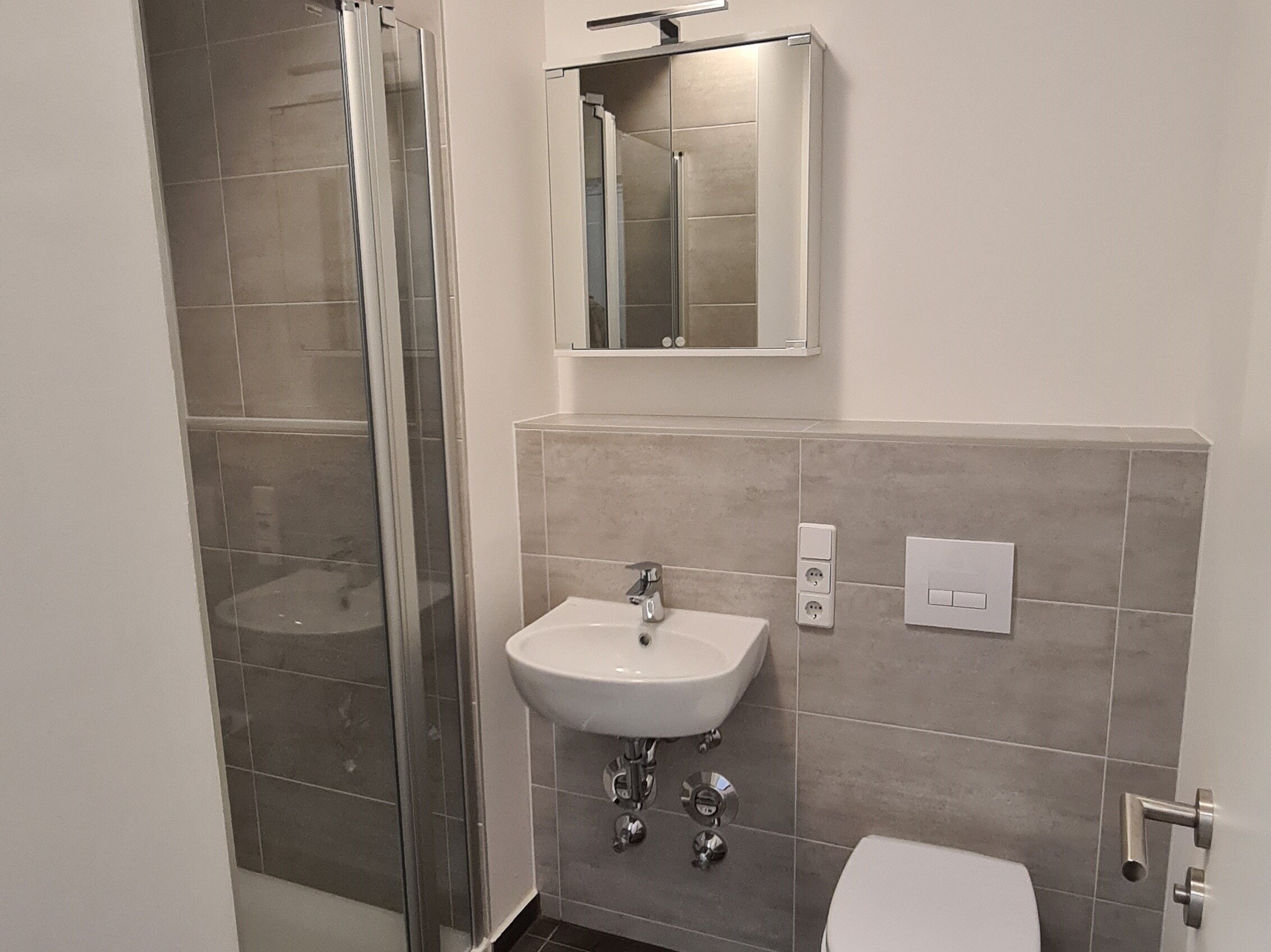 Modernes Badezimmer mit bodengleicher Dusche und Glasabtrennung, wandhängendem WC, kleinem Waschbecken mit Spiegel- und Lichtaufsatz sowie grau gefliesten Wänden und dunklem Boden.