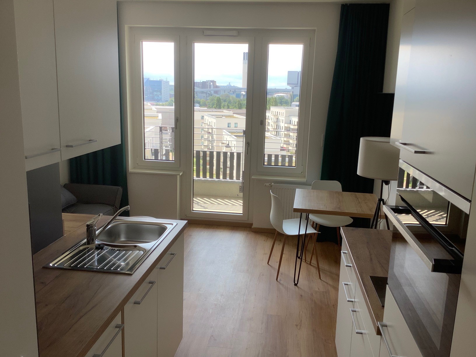 Kompakte Küche mit Spüle und Einbauschränken, Blick Richtung bodentiefe Fenster und Balkontür; vor dem Fenster stehen ein kleiner Tisch mit zwei Stühlen und eine Stehlampe, außerhalb sind weitere Wohngebäude zu sehen.